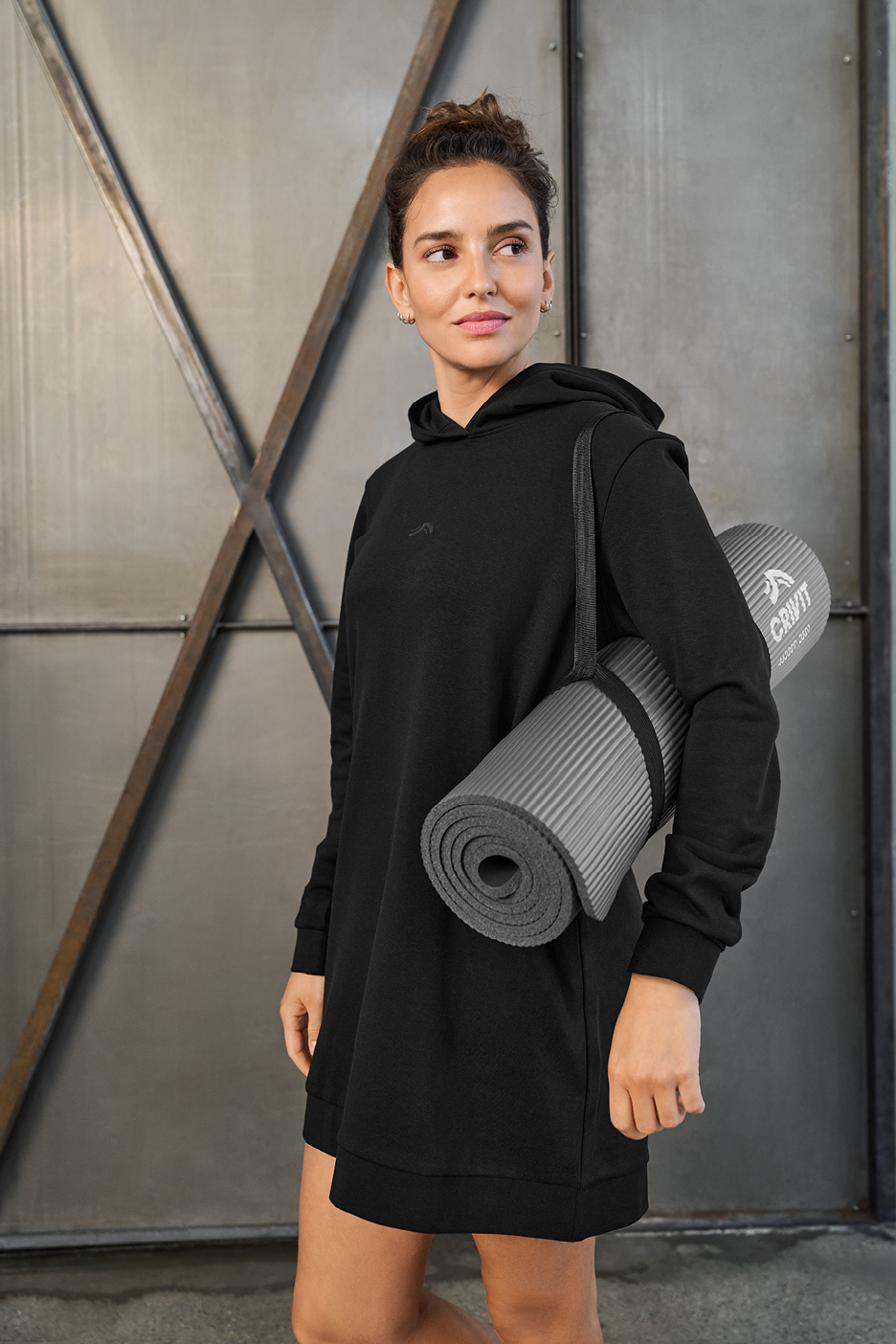 Žena u crnoj dukserici s kapuljačom nosi sivu prostirku za jogu preko ramena.