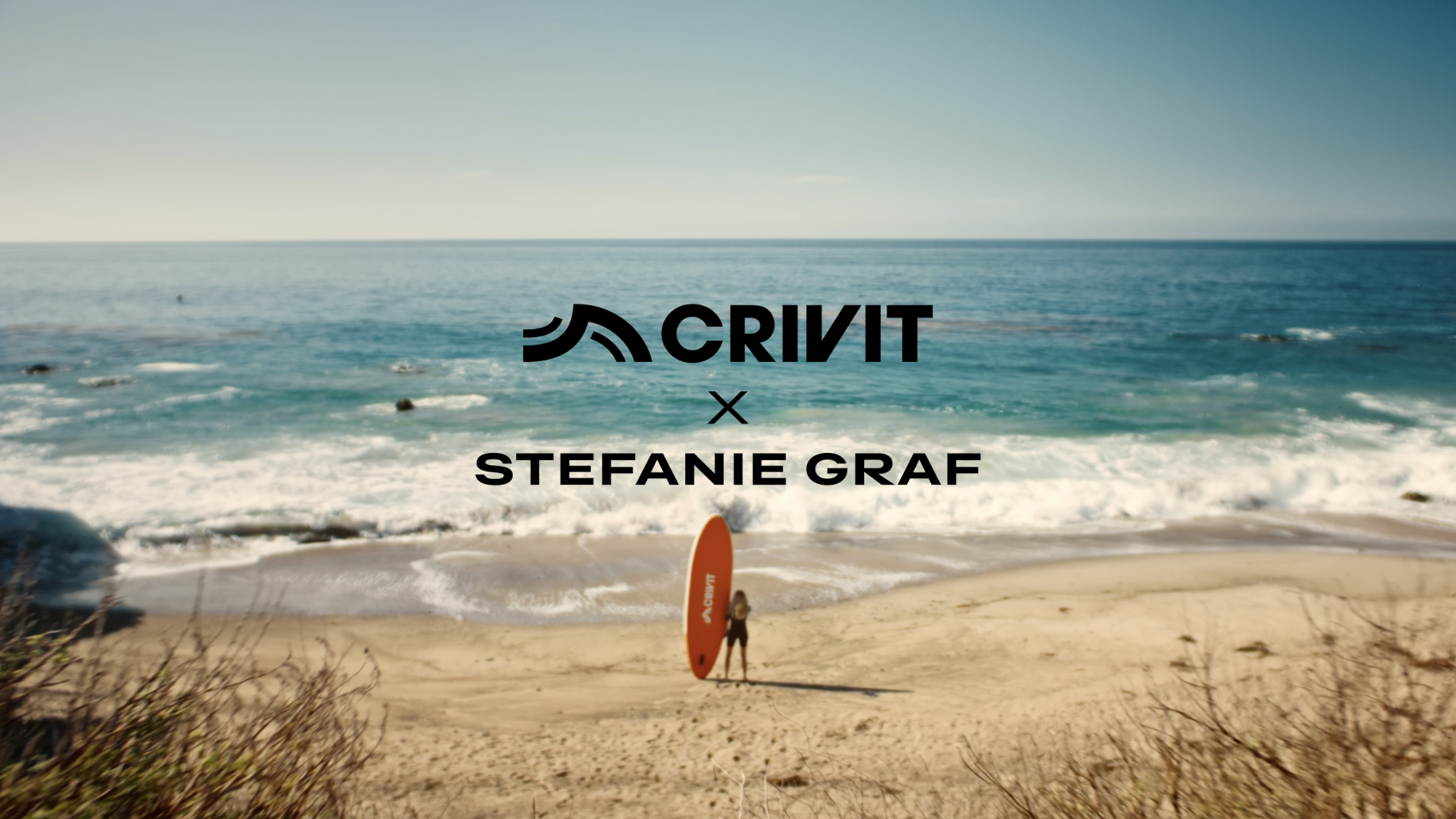 Žena s narančastom SUP daskom na pješčanoj plaži, s morem u pozadini.