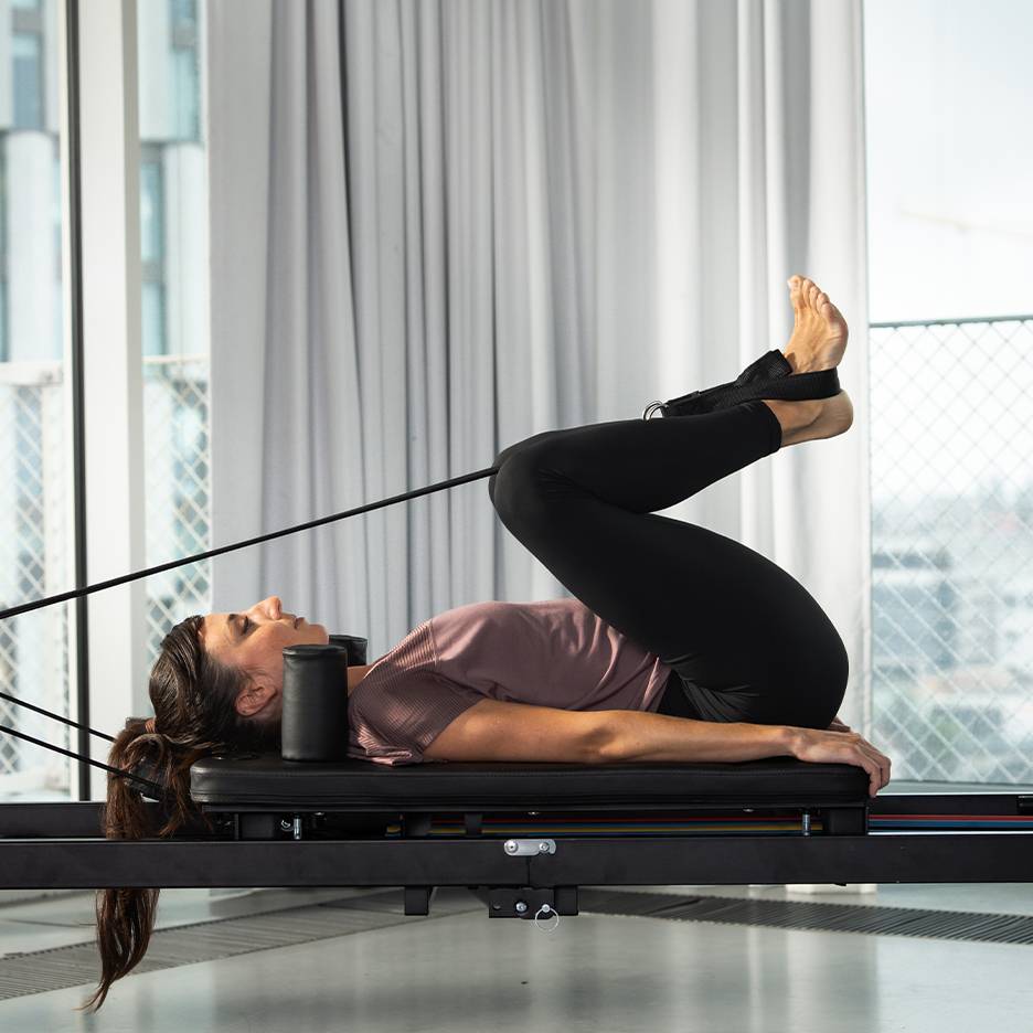 Žena vježba pilates na reformer spravi s podignutim nogama.