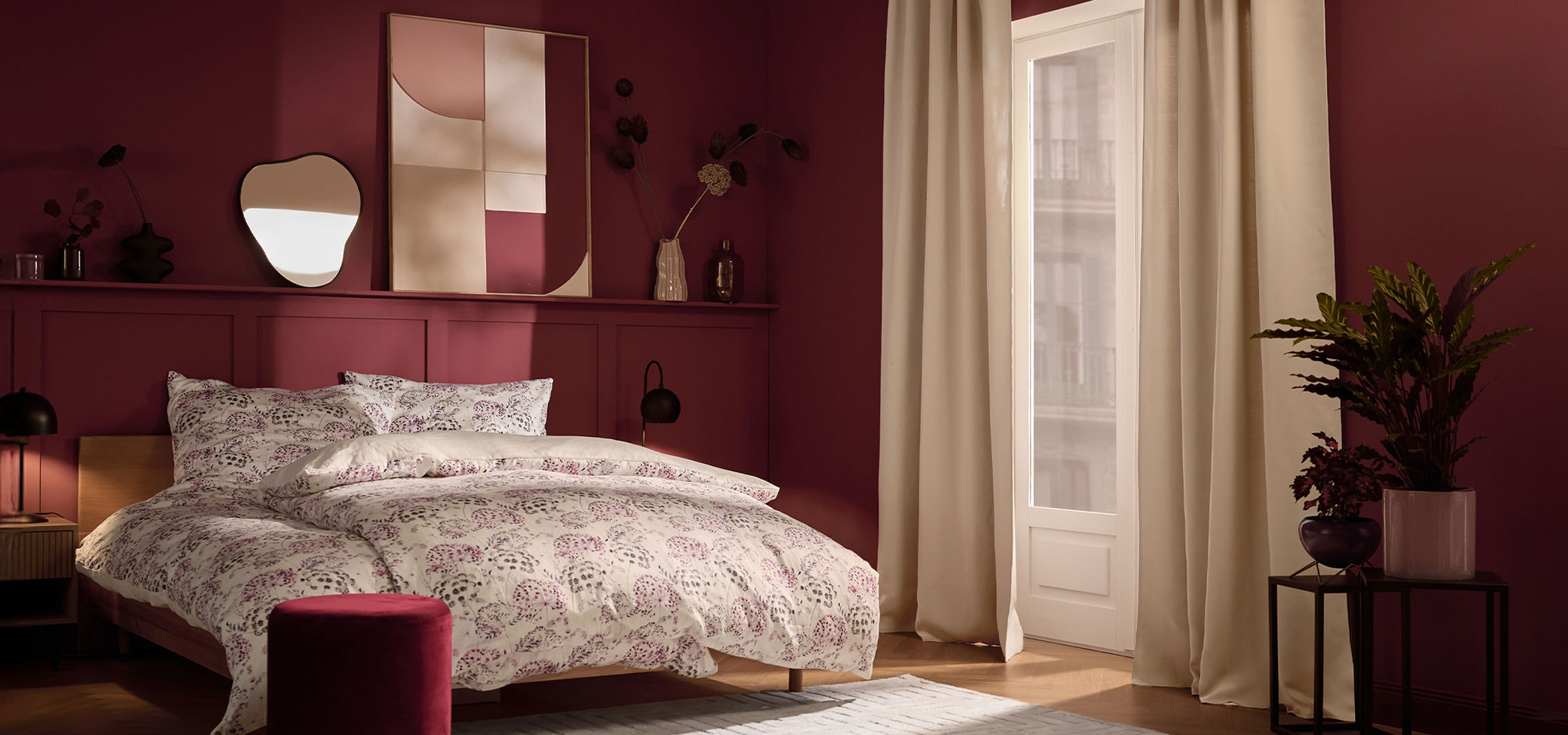 Spavaća soba s tamnocrvenim zidovima, krevetom s bijelom posteljinom s cvjetnim uzorkom i biljkama.