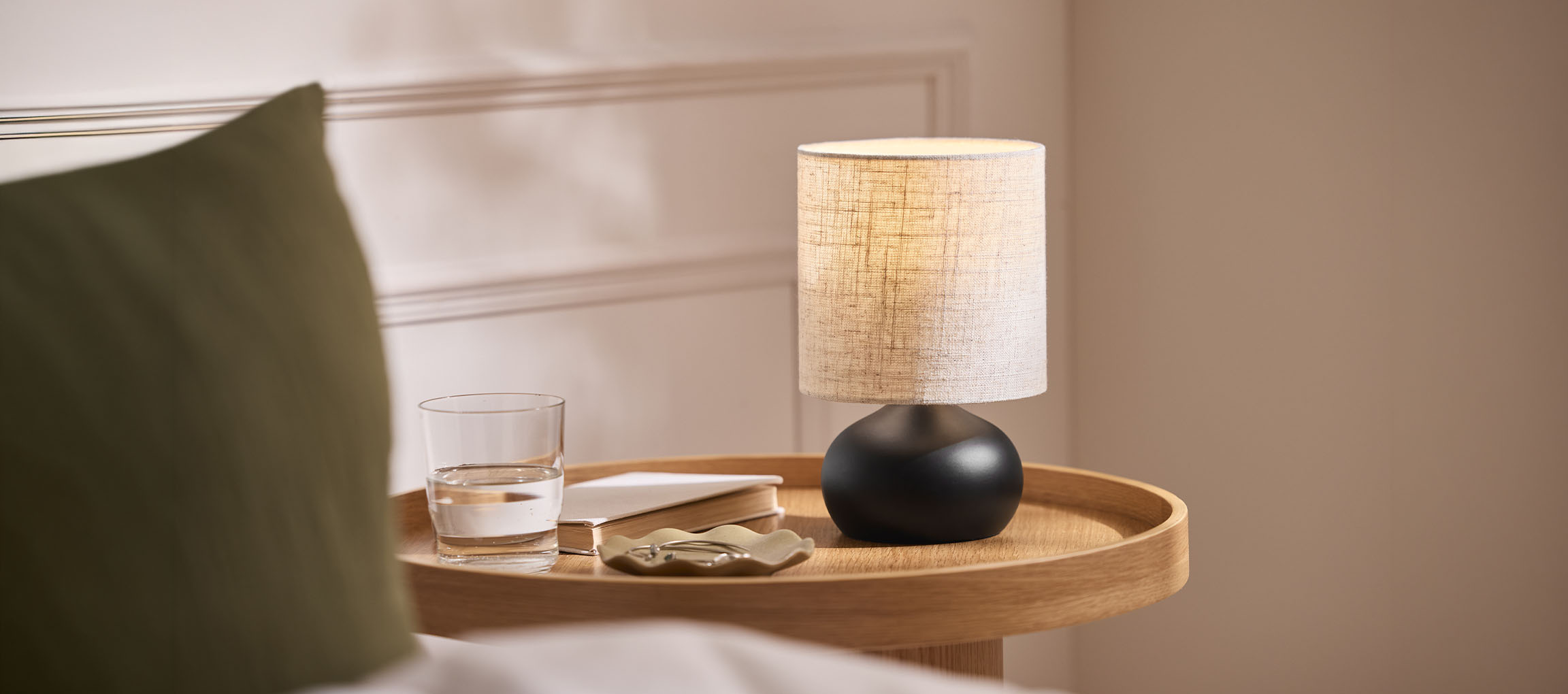 Noćna lampa s platnenim sjenilom, čaša vode i knjiga na drvenom pladnju.