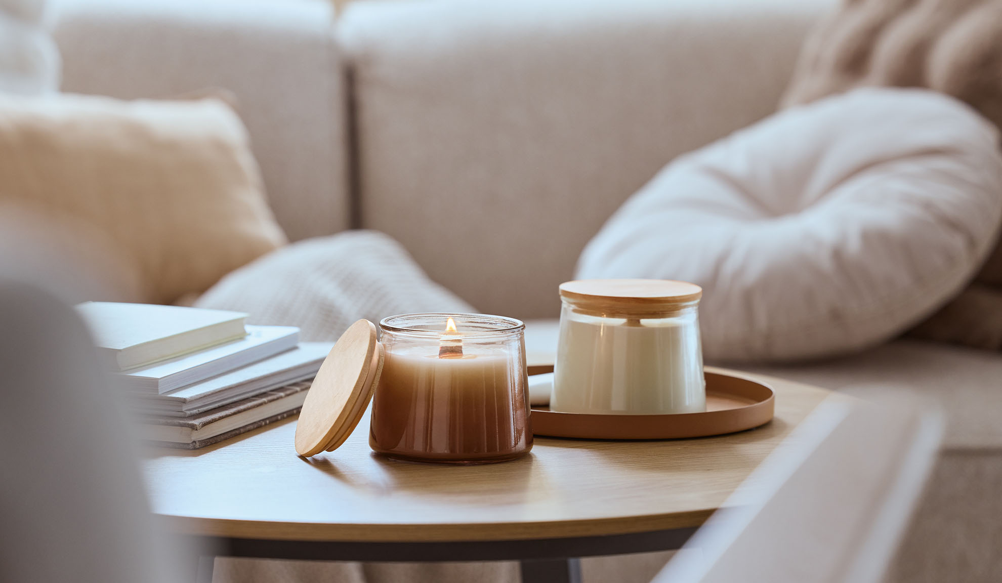 Dvije mirisne svijeće na drvenom stolu, jedna upaljena, s udobnom sofom u pozadini.