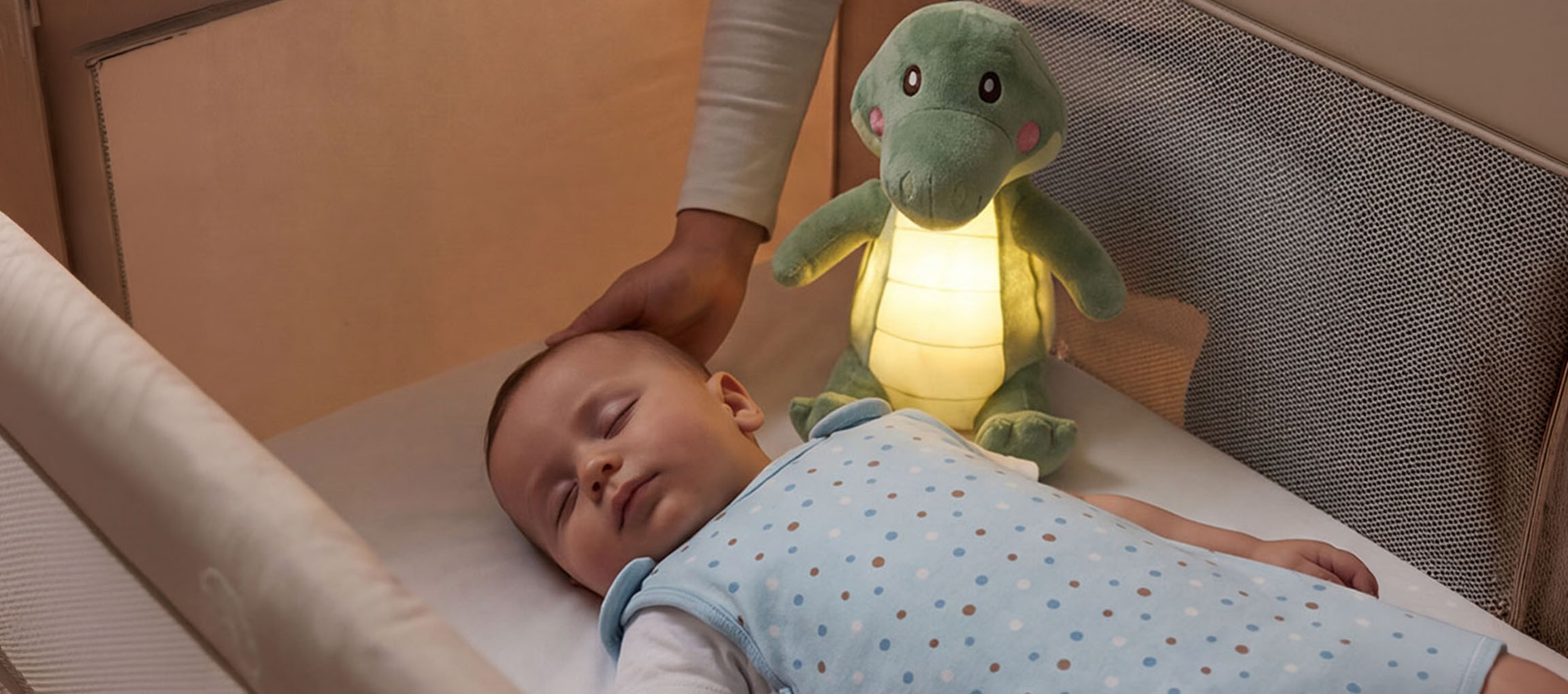 Beba spava u krevetiću s plišanom noćnom lampom, odrasla osoba mazi bebinu glavu.