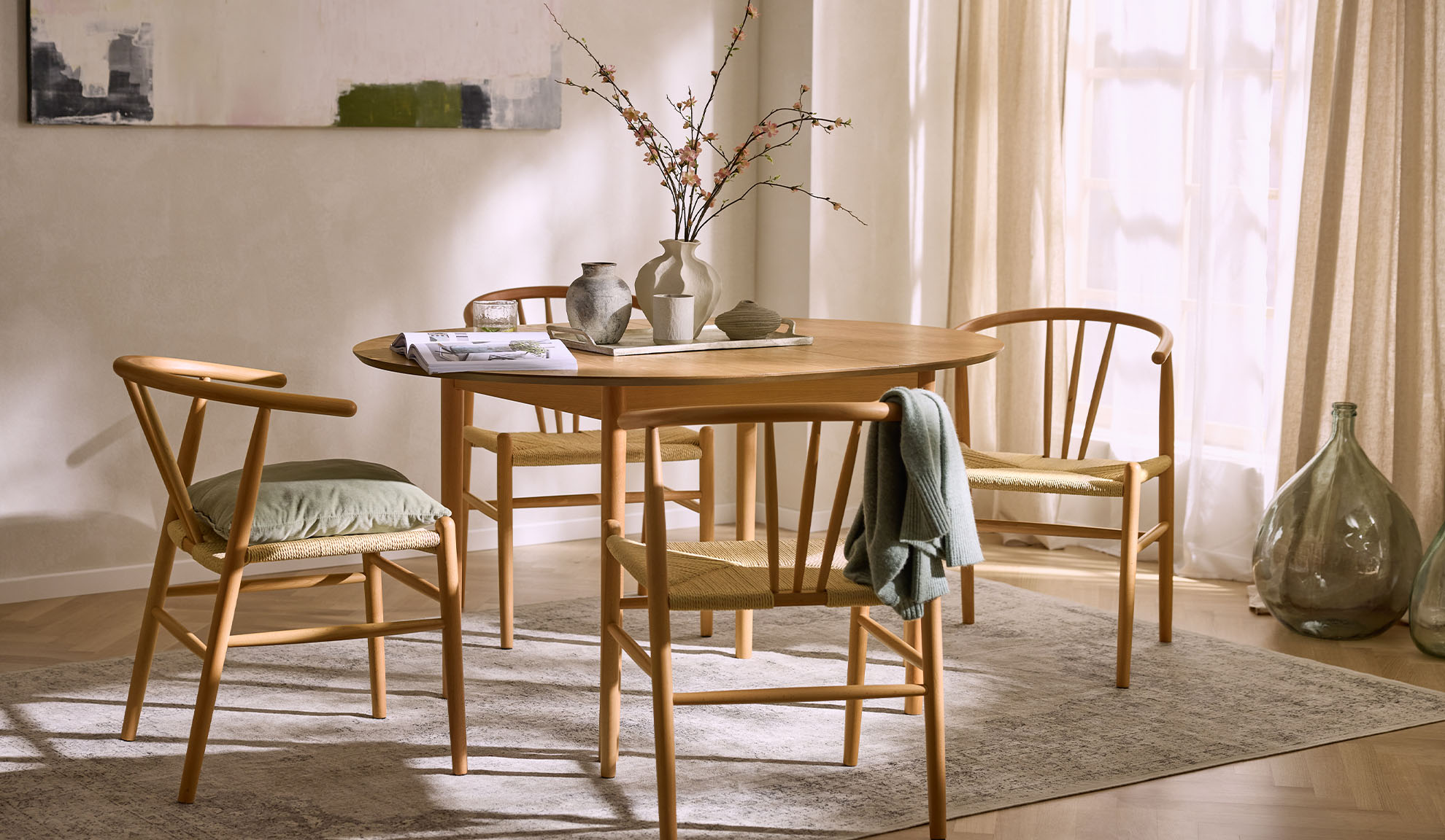 Svijetla blagovaonica s okruglim drvenim stolom, drvenim stolicama s pletenim sjedalima i cvjetnim dekorom.