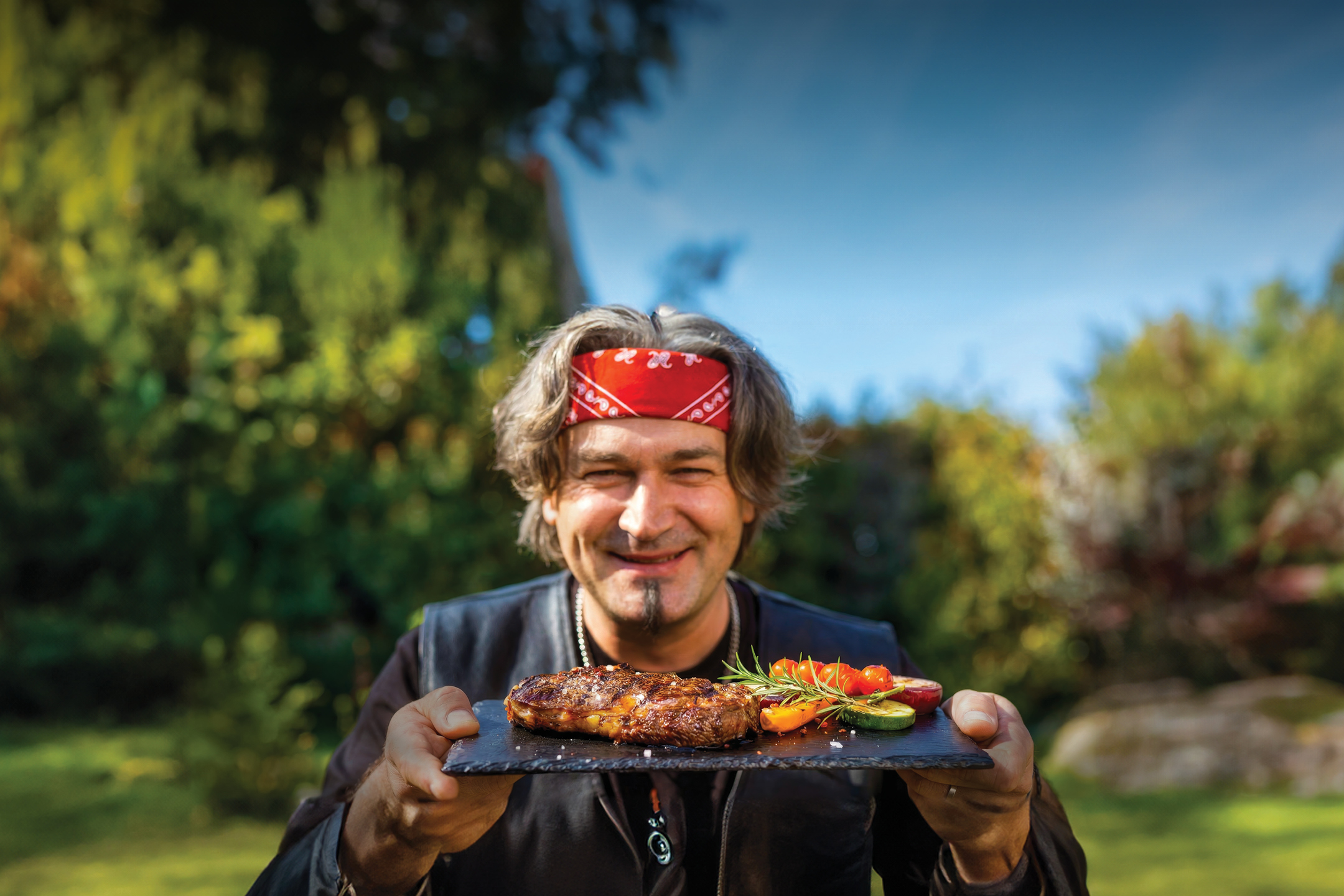 Muškarac s crvenom maramom drži pladanj s odreskom i povrćem na otvorenom.