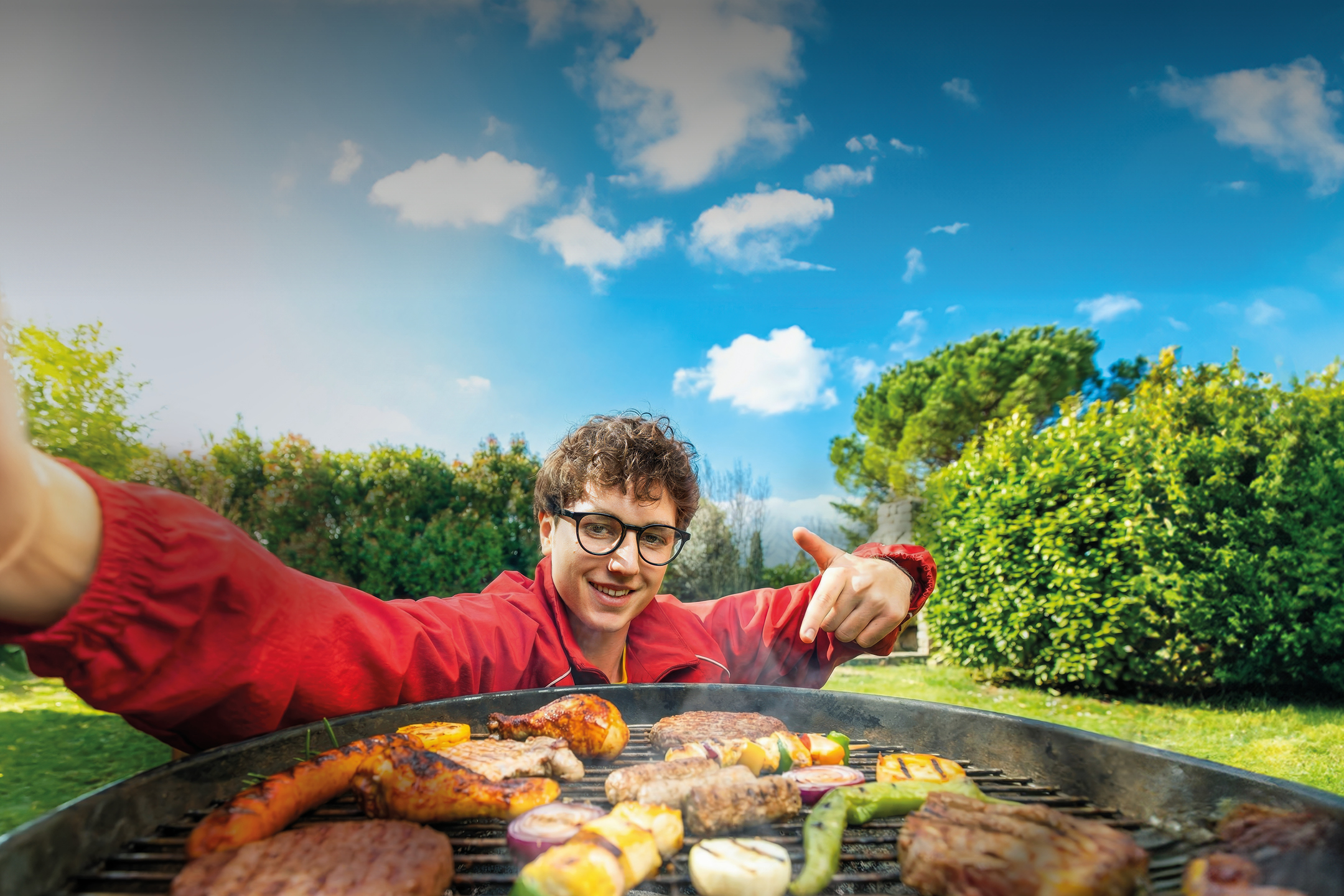 Mladić se smiješi dok pokazuje na roštilj pun mesa i povrća u vrtu.