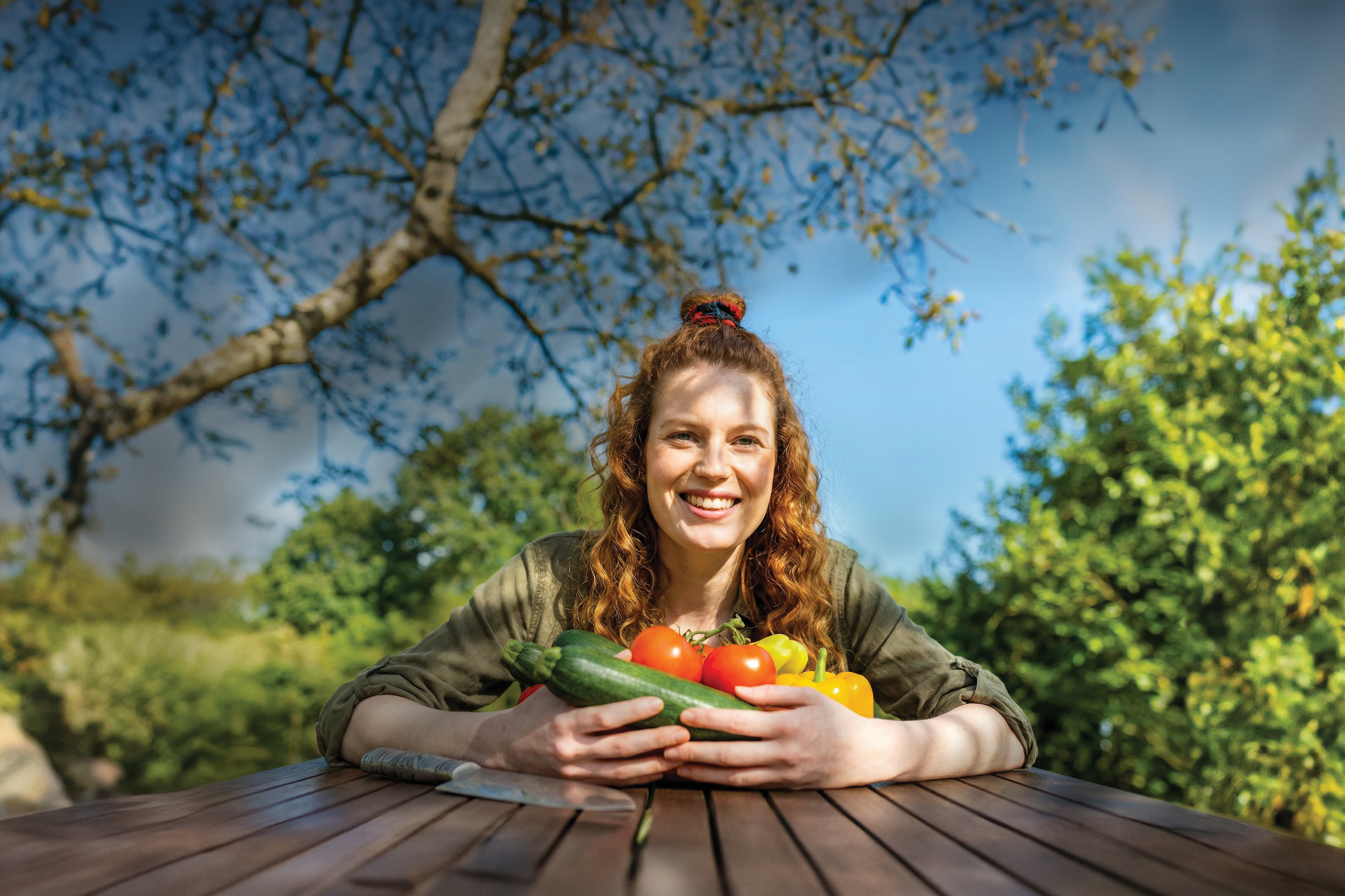 Žena s osmijehom drži tikvice, rajčice i paprike na drvenom stolu u vrtu.