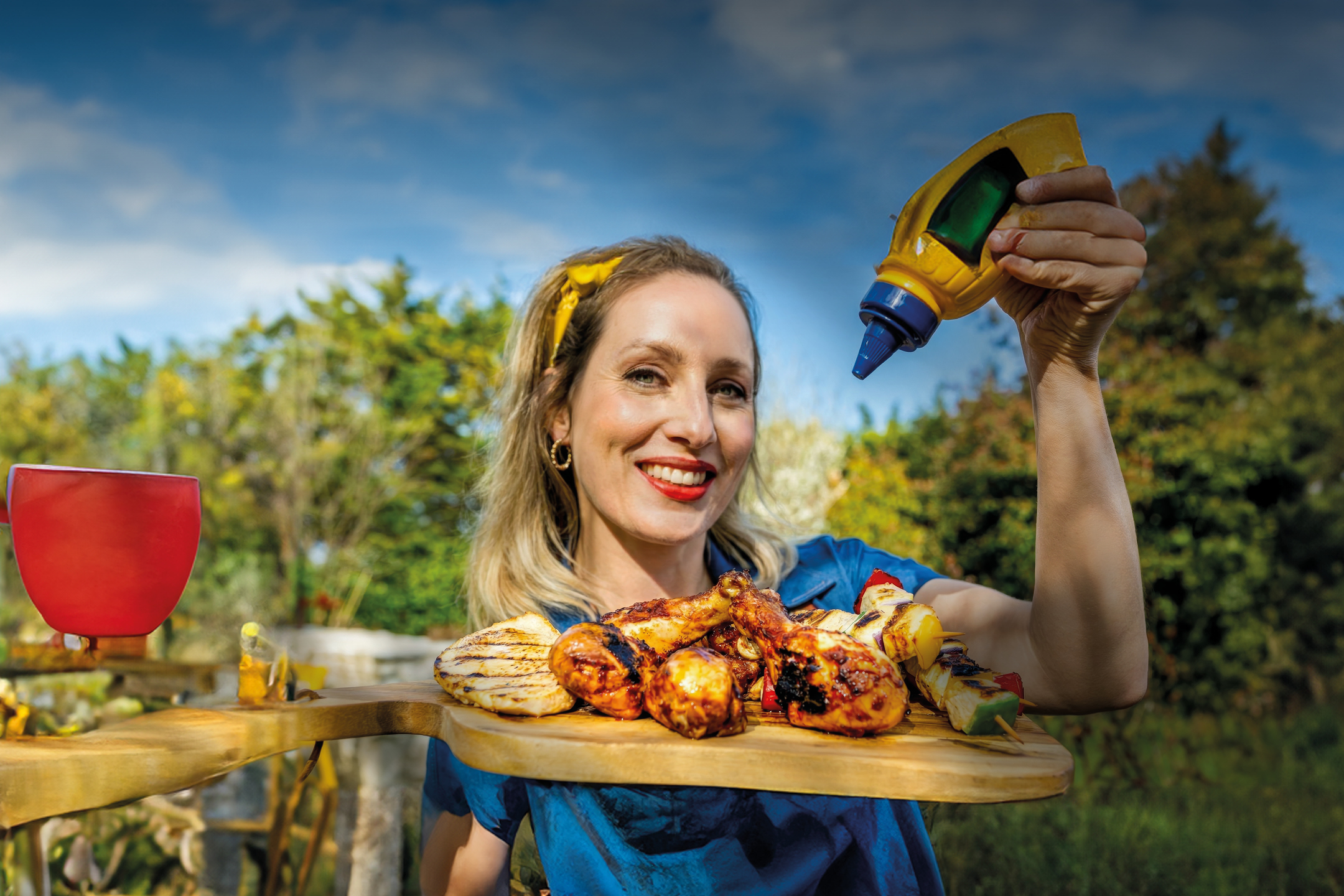 Žena drži pladanj s roštiljem i bocom senfa, smiješeći se u vrtu.