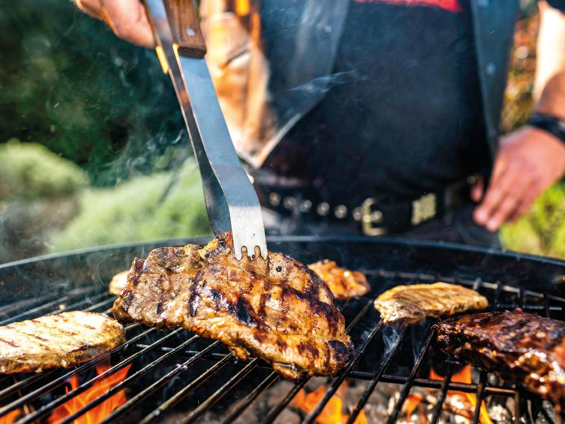 Muškarac okreće odreske na roštilju, s plamenom i dimom ispod rešetke.
