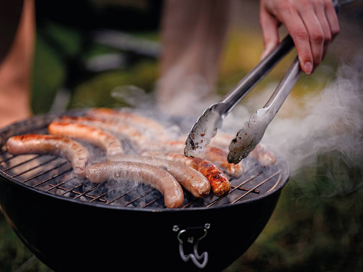 Kobasice na roštilju s dimom i rukom koja ih okreće hvataljkama.