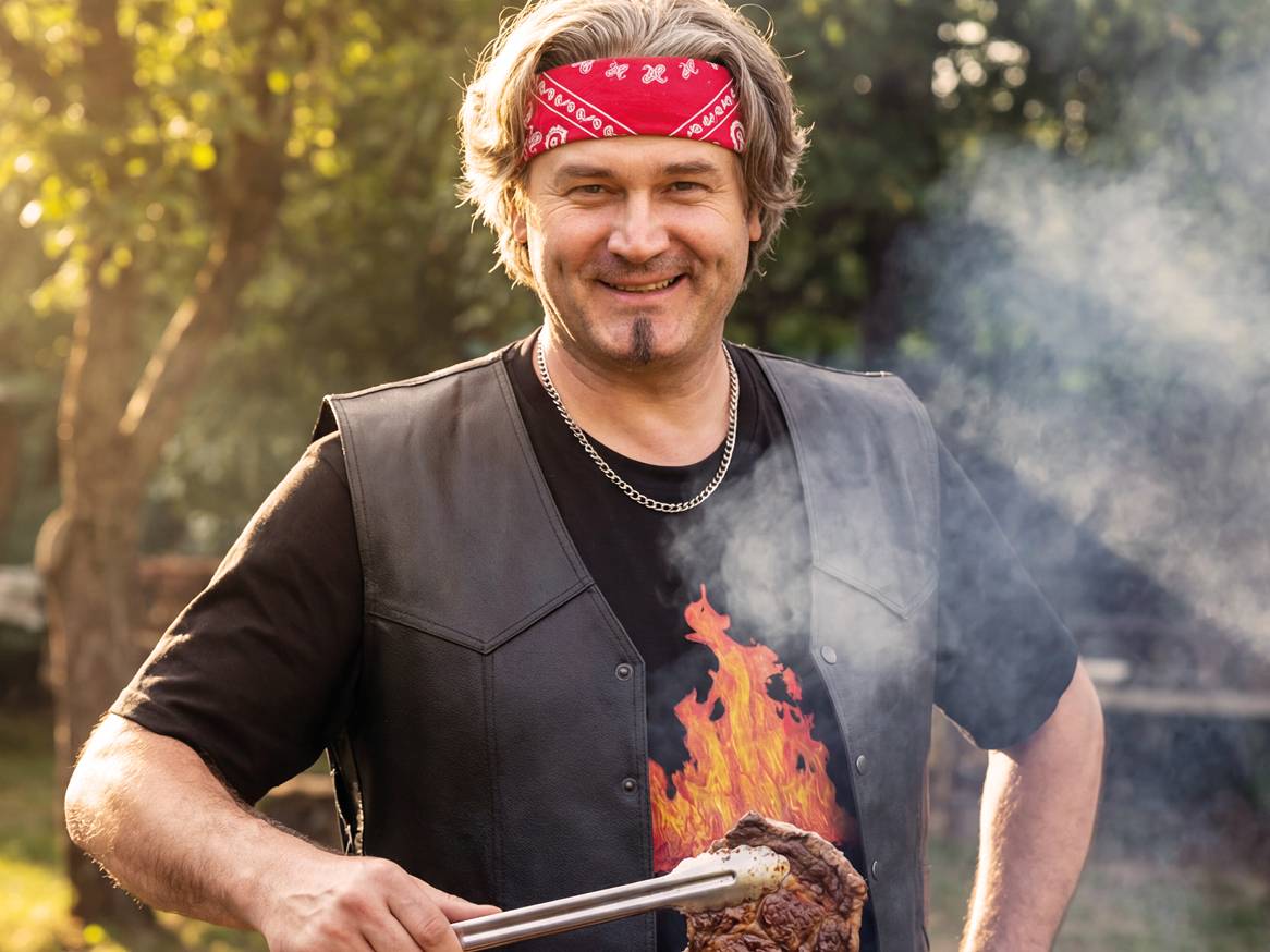 Muškarac u kožnom prsluku i majici s plamenom peče odrezak na roštilju.