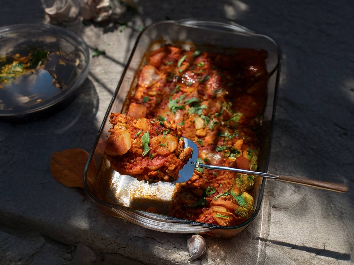 Zapečeno jelo od krumpira i povrća u staklenoj posudi za pečenje, s lopaticom koja vadi porciju.