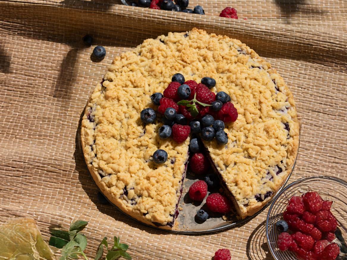 Pita s borovnicama i malinama na pletenoj prostirci, s kriškom izvađenom.