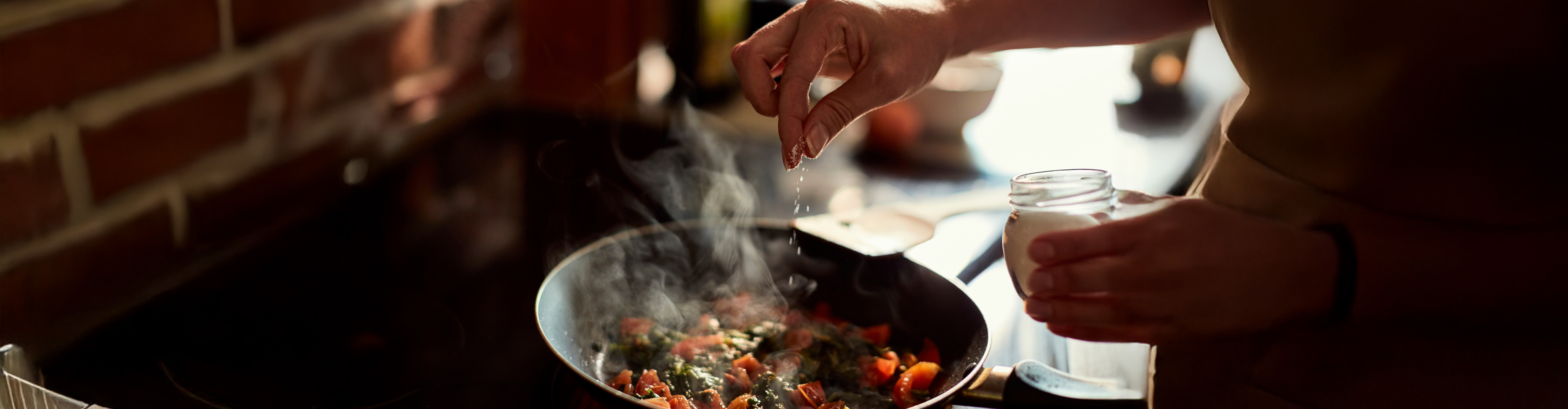 Ruka posipa začine u tavu s povrćem koje se kuha na štednjaku.