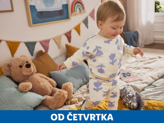 Beba u pidžami s uzorkom cvijeća stoji pored plišanog medvjedića i jastuka u dječjoj sobi.