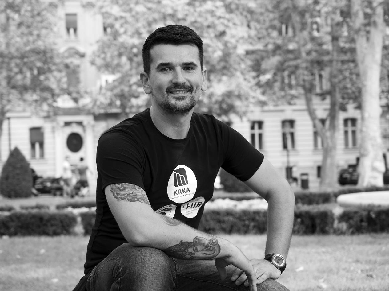 Muškarac u crnoj majici s logotipom Krka i tetovažama na rukama, sjedi vani.