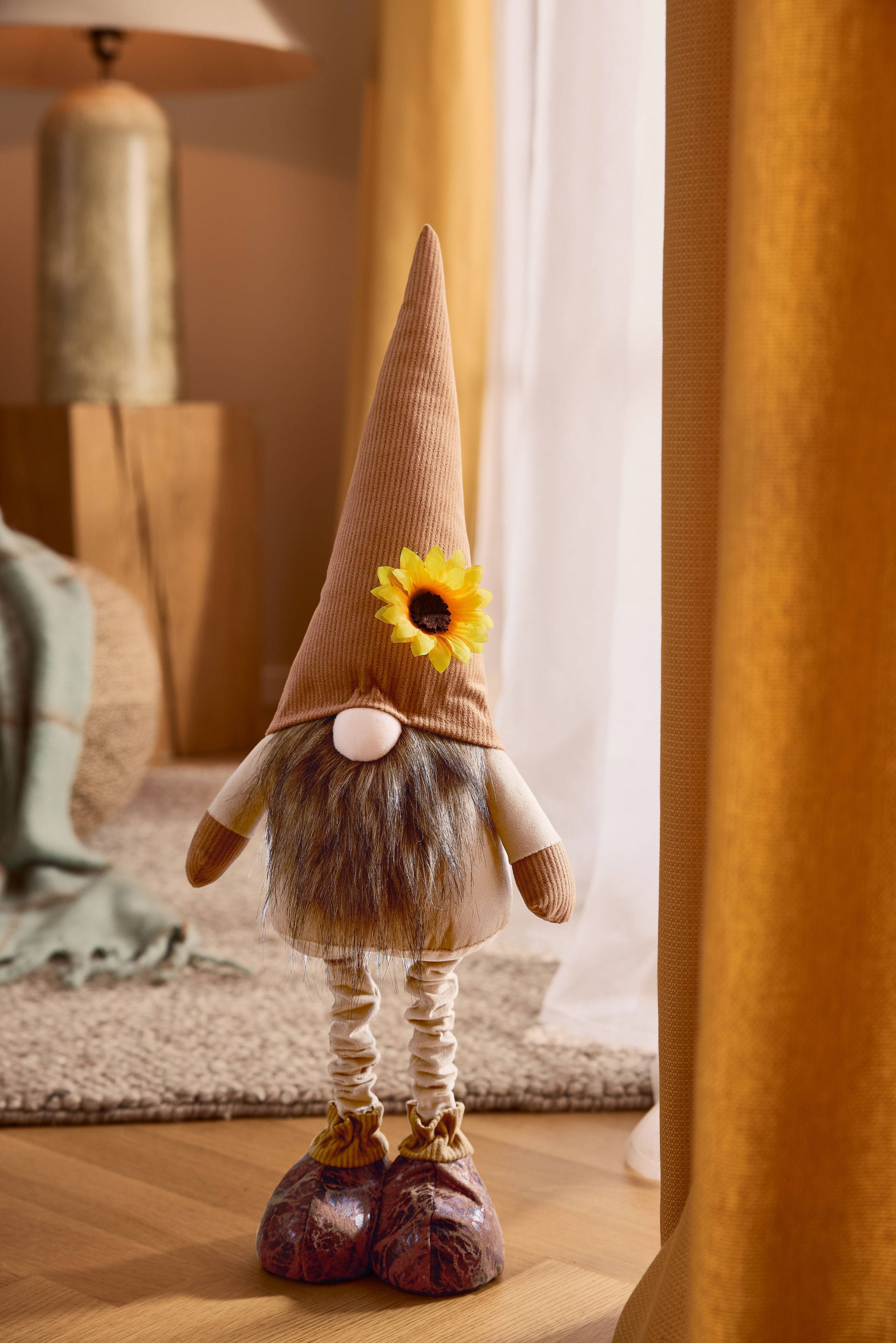 Plišani patuljak sa smeđom šiljastom kapom, bradom i suncokretom, u ugodnom okruženju.