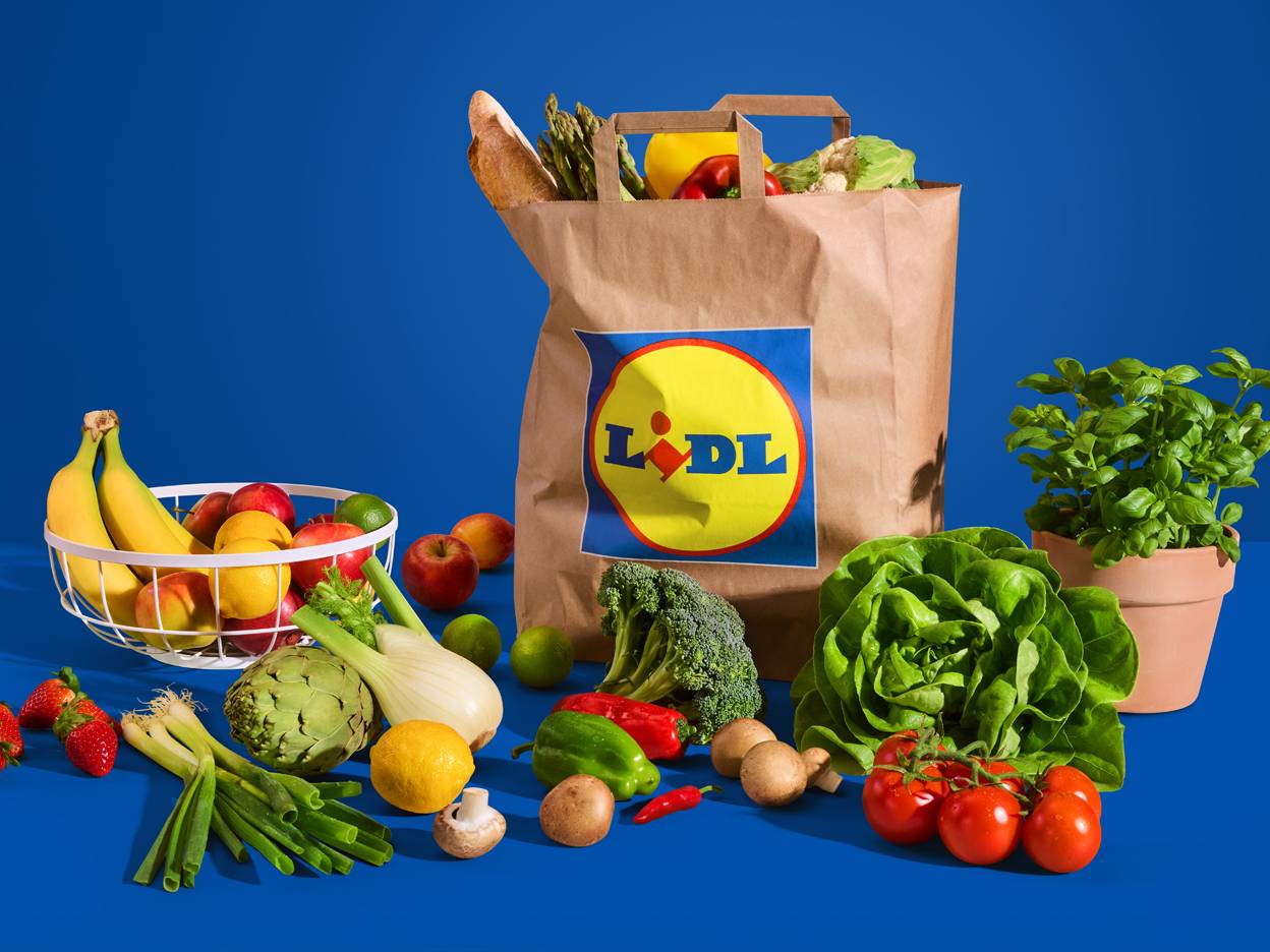 Smeđa papirnata vrećica s logotipom Lidla, okružena svježim voćem i povrćem na plavoj pozadini.