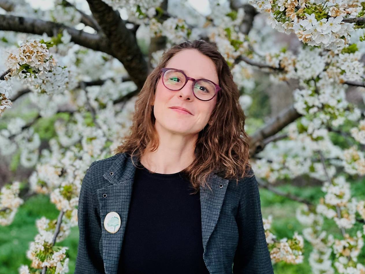 Žena s naočalama i brošem u obliku drveta, okružena bijelim cvjetovima.