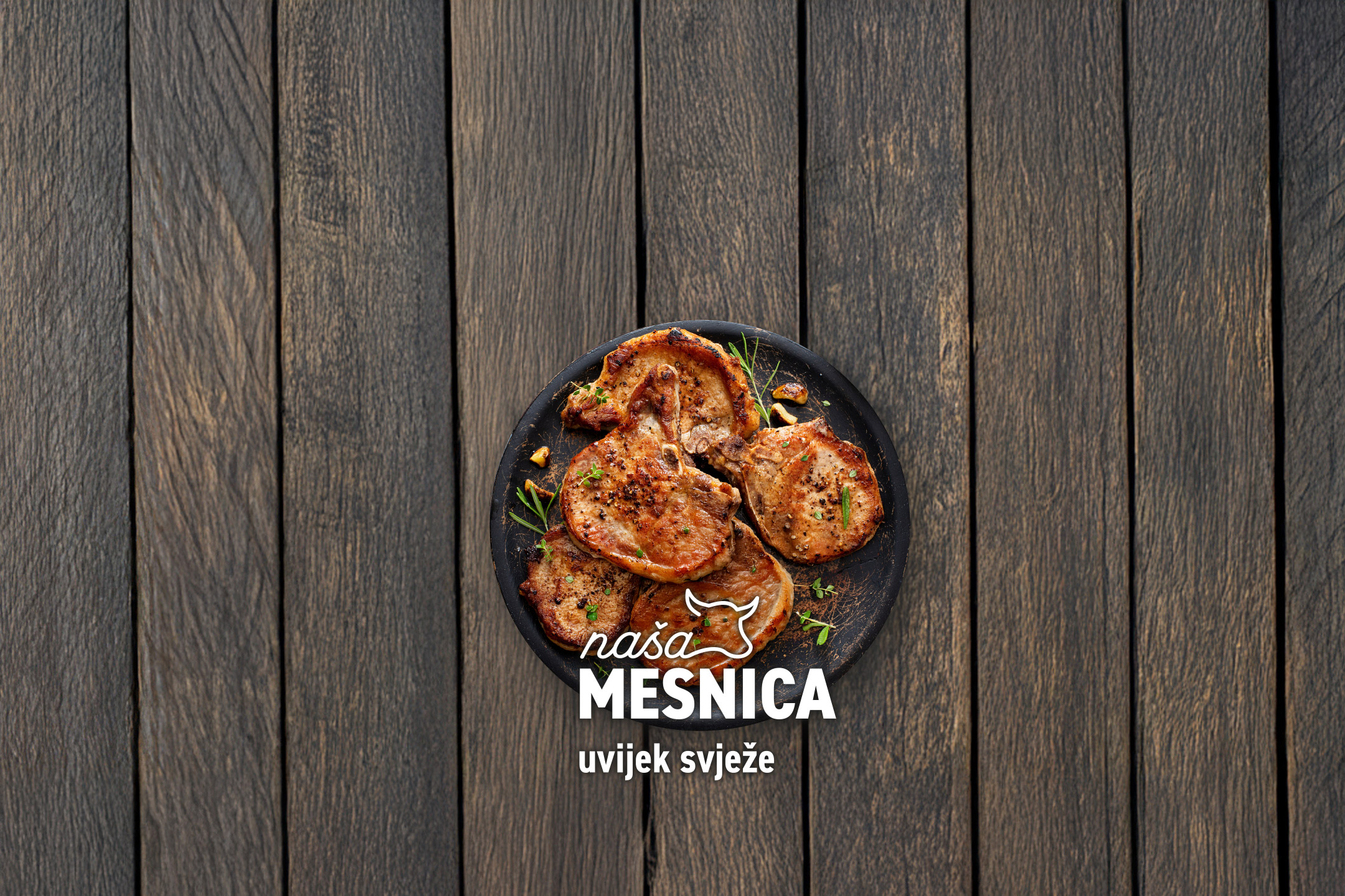 Pečeni svinjski kotleti s biljem na tamnom drvenom stolu, s natpisom 'Naša mesnica, uvijek svježe'.