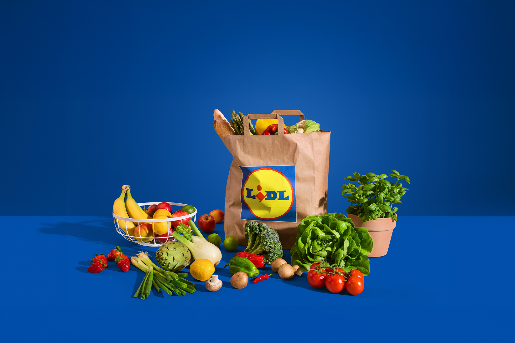 Lidl papirnata vrećica puna svježeg voća i povrća, uključujući salatu, rajčice, banane i jagode, na plavoj pozadini.