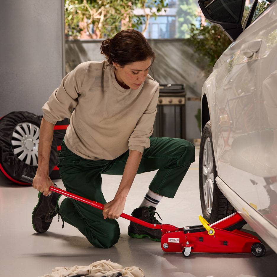 Žena koristi crvenu hidrauličnu dizalicu za podizanje bijelog automobila, s rezervnim kotačem u blizini.