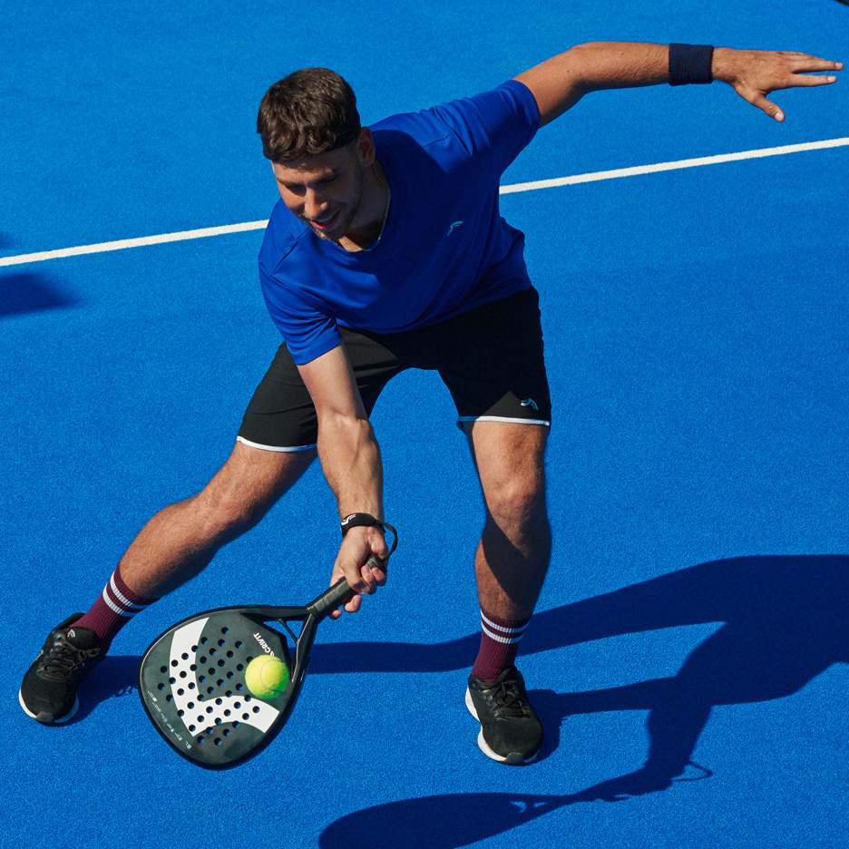 Muškarac u plavoj majici i crnim kratkim hlačama igra padel s reketom i lopticom.
