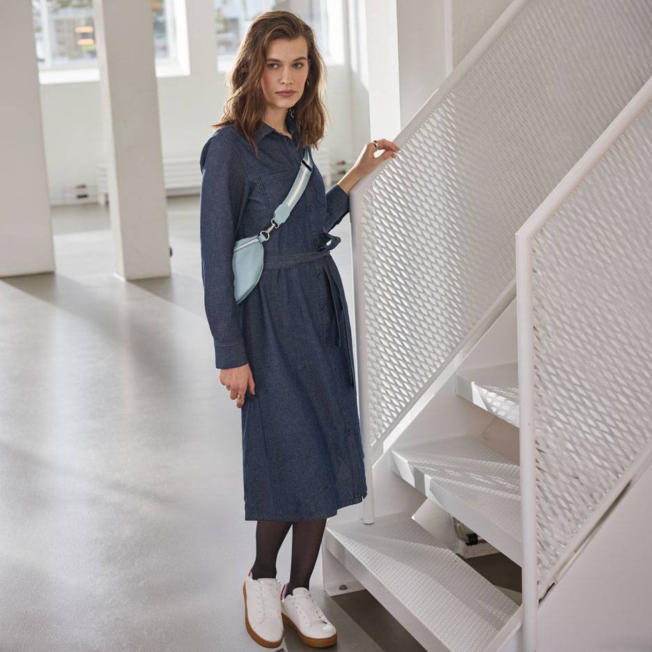 Žena u traper košulja-haljini, crnim najlonkama i bijelim tenisicama, sa svijetlo plavom torbicom oko struka.